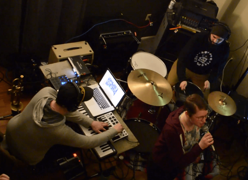 An overhead shot of three people performing in an electronic band with a drumset and keyboard and a vocalist.
